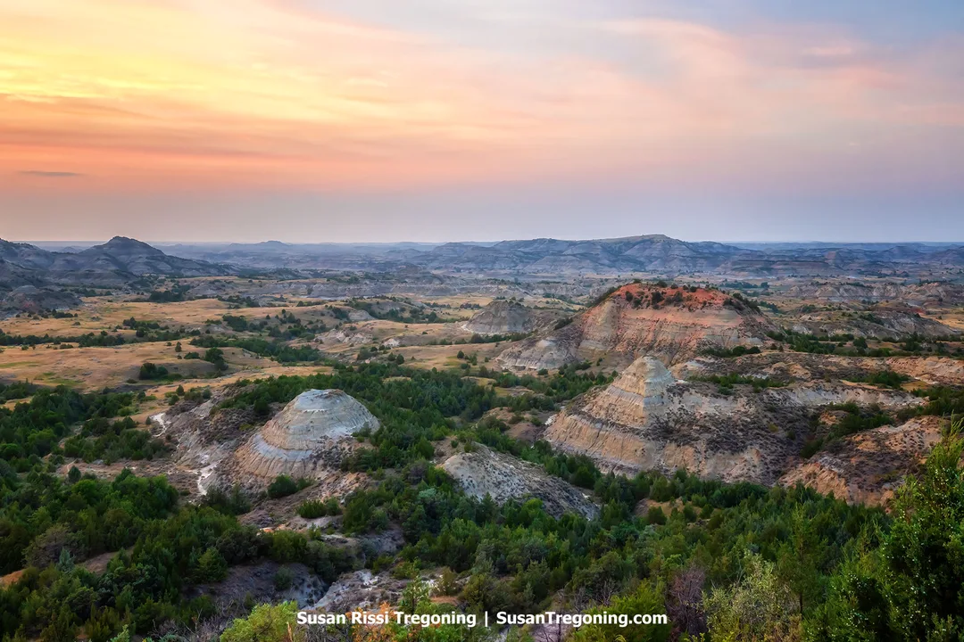 A wide sunset view over Painted Canyon in Theodore Roosevelt National Park, showing layered Badlands formations in shades of tan, red, and gray with scattered green vegetation. Soft orange and pink light fills the sky, highlighting the rugged hills and deep erosion patterns across the landscape.