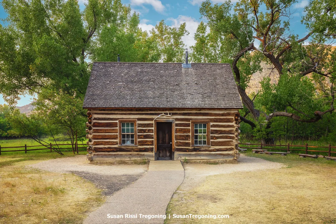 Theodore Roosevelt’s first home in the North Dakota Badlands — the historic Maltese Cross Cabin at the South Unit Visitor Center of Theodore Roosevelt National Park. The log cabin stands amid tall green trees and open prairie under a bright blue sky.
