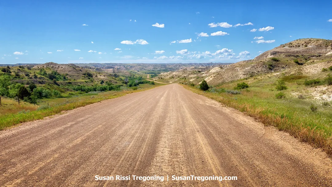 A gravel and dirt road winds through the rolling hills of the North Dakota Badlands under a clear blue sky. This remote route leads to the Elkhorn Ranch Unit of Theodore Roosevelt National Park, where the final three miles often require a four‑wheel‑drive and/or high‑clearance vehicle to navigate the rugged terrain.