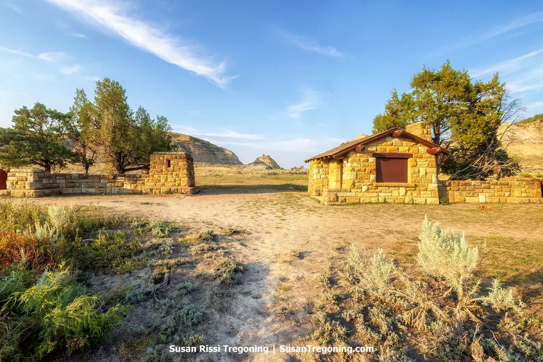 Historic sandstone entrance building and gate at the Old East Entrance of Theodore Roosevelt National Park’s South Unit, built by the Civilian Conservation Corps in 1938. The structure stands amid open prairie with scattered junipers and distant badlands formations under a clear blue sky.