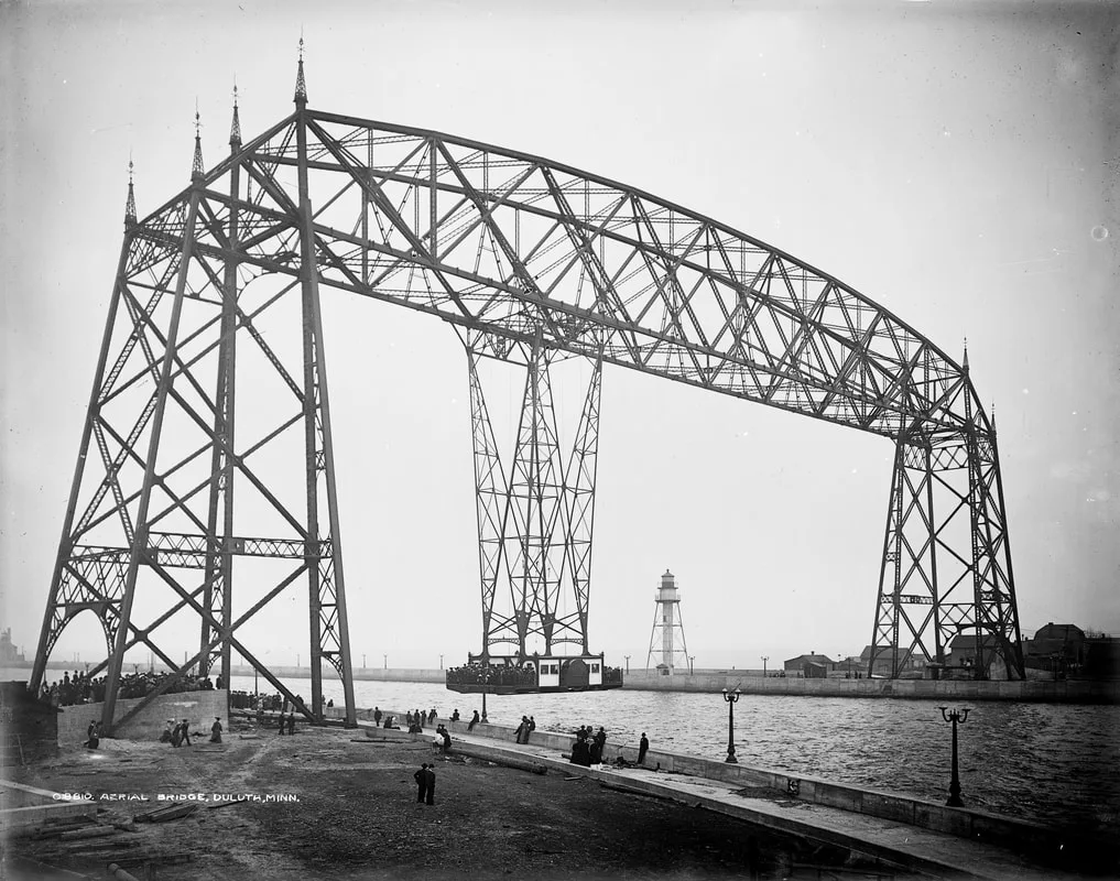 A historic view of Duluth’s original Aerial Bridge shows its steel truss structure with the suspended gondola car positioned beneath it. People stand near the base of the bridge along the waterfront, and a lighthouse and several buildings are visible in the background under a clear sky.