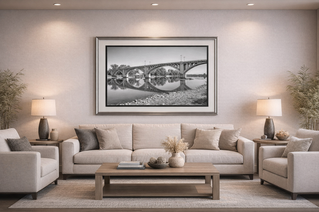 A warmly lit living room with a large beige sofa centered beneath a framed black‑and‑white photograph of the Lincoln Memorial Bridge in Vincennes, Indiana, reflected in calm water. Matching beige armchairs sit on either side of the sofa, with wooden end tables and identical table lamps beside them. A wooden coffee table holds a vase of dried flowers and decorative objects. Light-colored walls, recessed ceiling lights, and tall potted plants in the corners create a balanced, serene space.