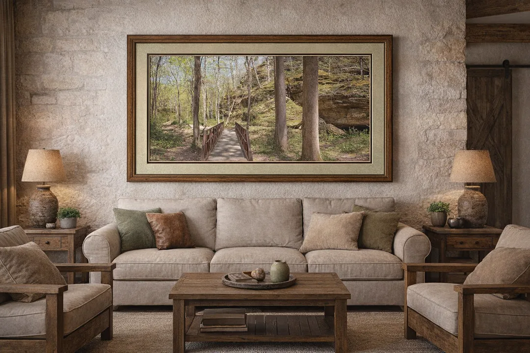 A rustic living room with a beige sofa and matching armchairs, centered beneath a framed panoramic photograph of a forest scene from Ferne Clyffe State Park. The artwork shows a metal-railed footbridge leading into a wooded area with tall trees and a moss-covered rock formation. The photograph is double-matted with a light greenish-beige top mat and a wider dark brown bottom mat, framed in warm wood. It hangs five inches above the sofa, leaving four inches of space on either side. The room features a stone wall, wooden coffee table, and earthy-toned décor.