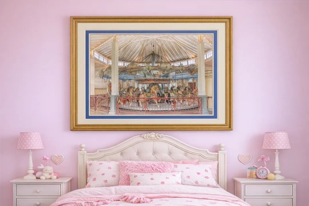 A framed photograph of a vintage carousel hangs above a white tufted bed in a little girl’s bedroom with soft pink walls. The image is double‑matted with a cream outer mat and a wider blue inner mat, all enclosed in an ornate gold frame. The bed is dressed in pink and white bedding with decorative pillows, and flanked by white nightstands holding a pink lamp, plush bunny, and pastel accessories.