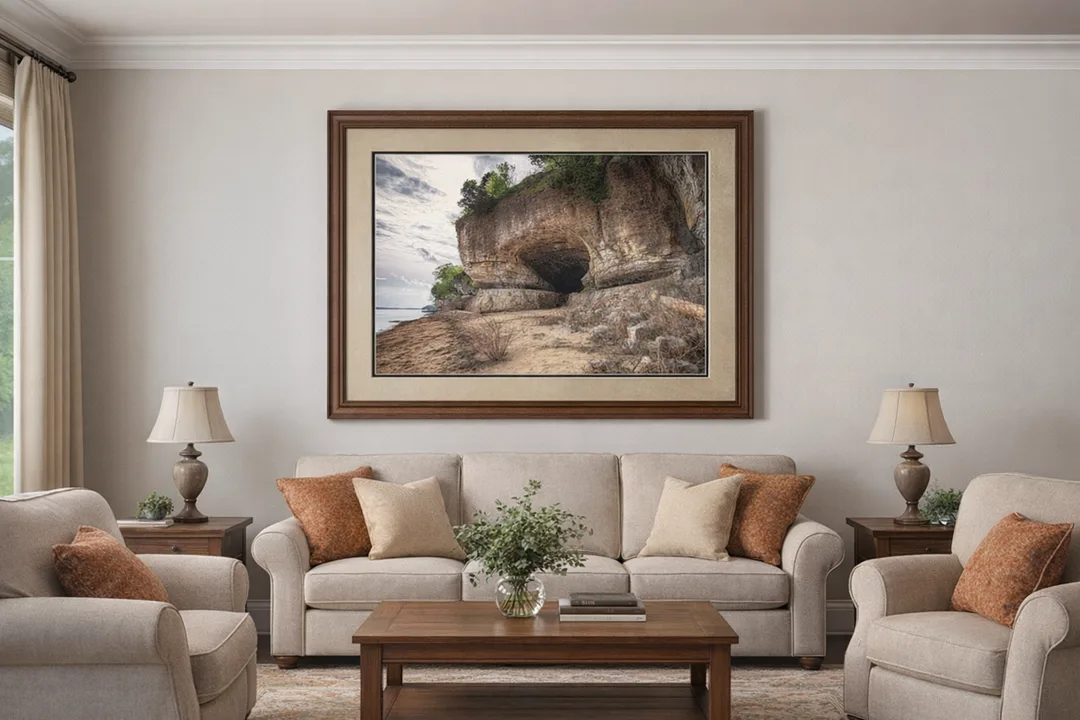 Framed landscape photograph of Cave‑In‑Rock hangs above a beige sofa in a comfortable living room. The artwork features a limestone cave with a sandy shoreline, double-matted with a light beige top mat and dark brown bottom mat, and set in a walnut wood frame. Matching beige armchairs, warm-toned throw pillows, and wooden furniture complete the cozy, coordinated space.