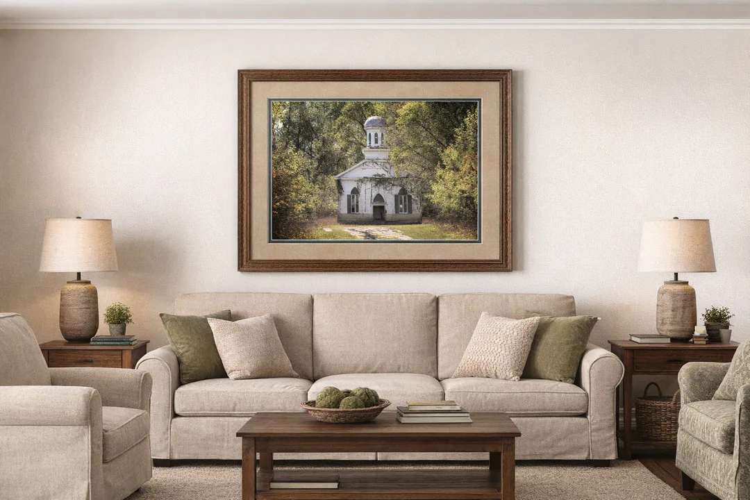 A framed photograph of an old white wooden church with a domed cupola hangs on a wall above a beige sofa in a cozy living room. The artwork is double‑matted with a light top mat and a darker bottom mat inside a wood frame. The room includes neutral‑toned furniture, throw pillows, wooden side tables with lamps, a coffee table with books and décor, and soft natural lighting.
