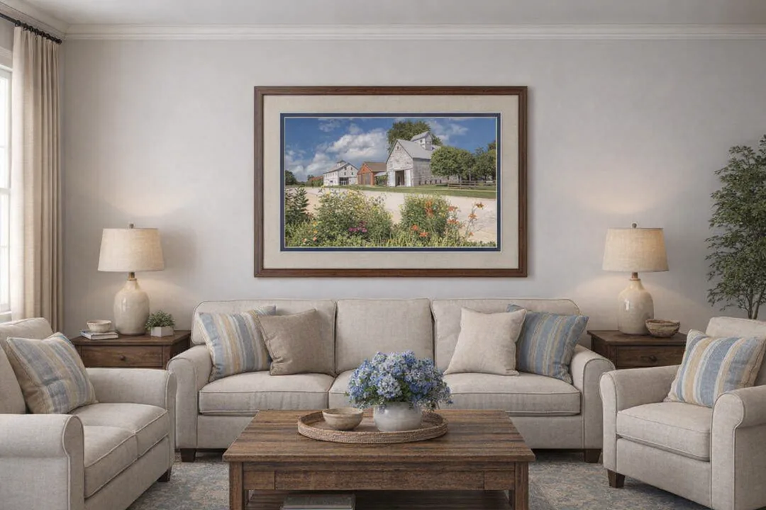 Framed farm photograph displayed in a cozy living room. The image features barns and colorful garden flowers under a bright sky, set in a wood frame with two mats: a light tan top mat and a darker blue bottom mat. The artwork hangs centered above a beige sofa, flanked by matching armchairs and rustic wood furniture. Blue hydrangeas, striped cushions, and soft lighting complete the warm, farmhouse-style setting, creating an inviting showcase for the wall art.