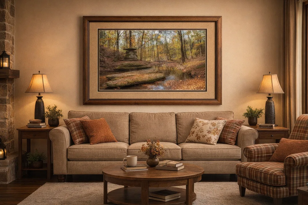 A large autumn landscape photograph hangs above a beige couch in a rustic living room. The image shows a shallow creek in a fall forest with yellow and orange foliage and a layered rock formation beside the water. The room includes plaid and leaf‑patterned pillows, warm lighting, wooden furniture, and a cozy, homey atmosphere.