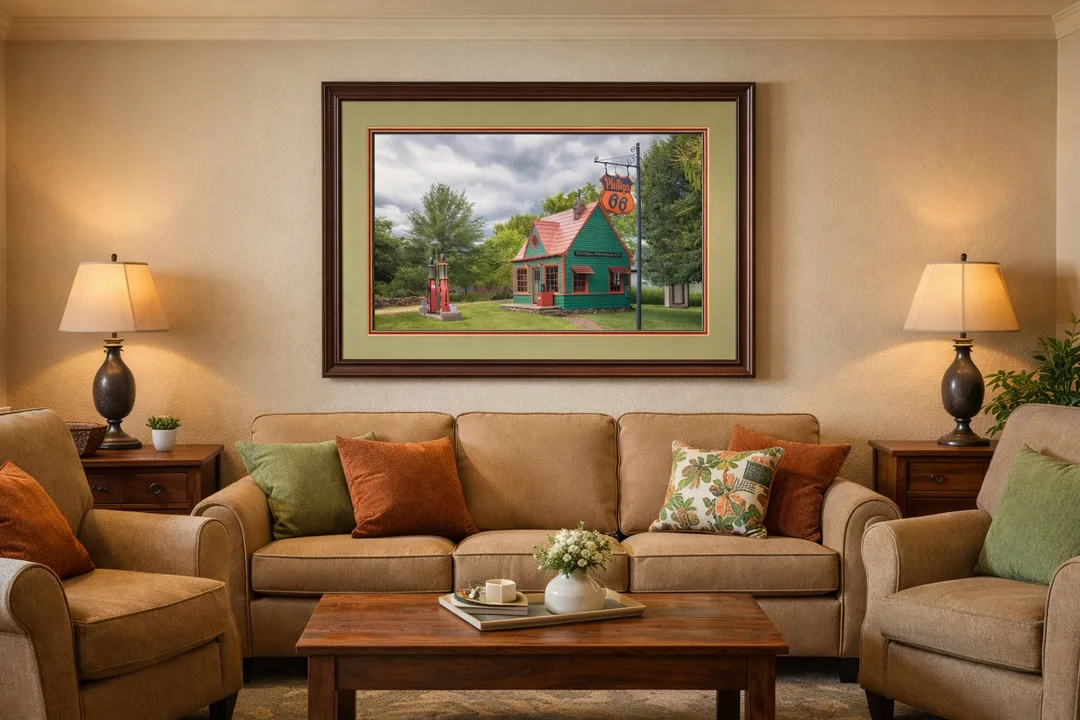   
A framed artwork hangs above a beige sofa in a living room, showing a small green building with a red roof, two vintage red gas pumps, and a Phillips 66 sign set in a grassy landscape under cloudy skies.