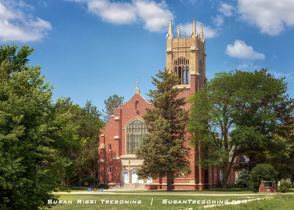A large brick Catholic church with a tall central tower and pointed‑arch windows stands among trees under a bright blue, partly cloudy sky.
