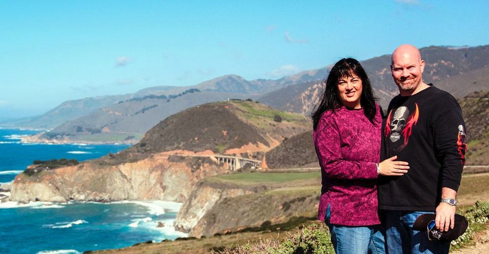 Susan and Paul Tregoning stand together at a scenic overlook above the iconic Bixby Bridge on California’s Big Sur coastline, with rugged cliffs, a sweeping concrete arch, and waves breaking along the shore below. Susan wears a purple sweater and Paul wears a black shirt with a skull‑and‑flame graphic. Clear blue sky and distant mountains frame the dramatic coastal landscape.