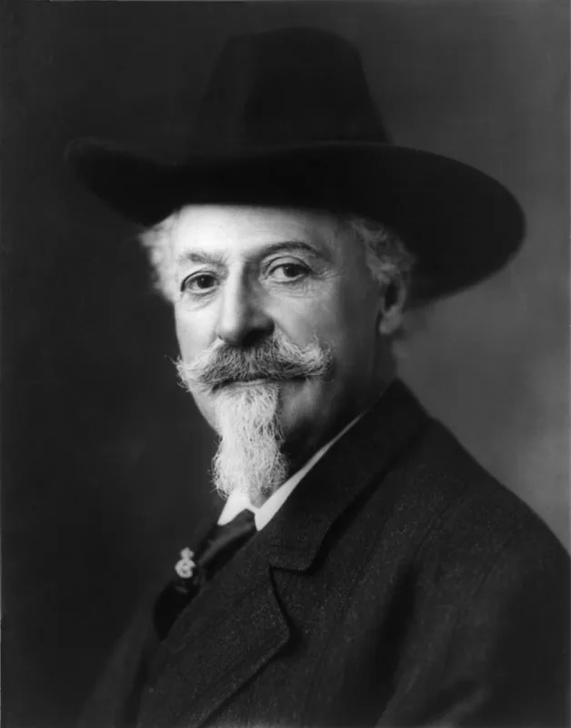 A black‑and‑white studio portrait of William F. Cody, shown with a wide‑brimmed hat, long hair, and a curled mustache and goatee, wearing a dark jacket, white shirt, and tie against a plain backdrop.