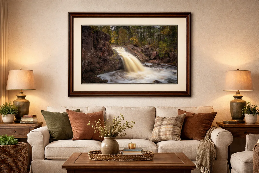 A warm, inviting living room features a large framed waterfall photograph as the focal point above a neutral sofa. The artwork is double-matted in complementary earth tones and set in a rich wood frame, enhancing the golden hues of the cascading water and autumn forest. Soft lighting, textured pillows, and rustic wood furniture echo the natural palette of the piece, creating a cozy, harmonious space that highlights the art as a statement centerpiece.